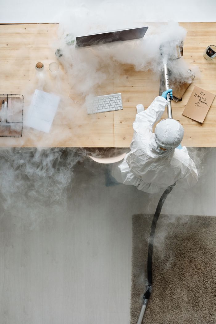 A person in protective gear sanitizing an office desk using fumigation equipment during daytime.